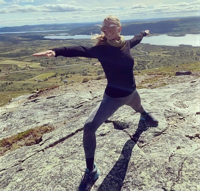 En kvinne som gjør en yogastilling på en fjelltopp med utsikt over et landskap med vann og fjell.